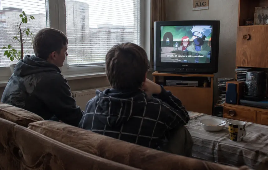 Lithuanian teenagers watching a dubbed anime show on television in a modest apartment during the early 2010s.