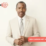 Ben Carson Net Worth: man in tan suit with flag pin, hands clasped, cream backdrop
