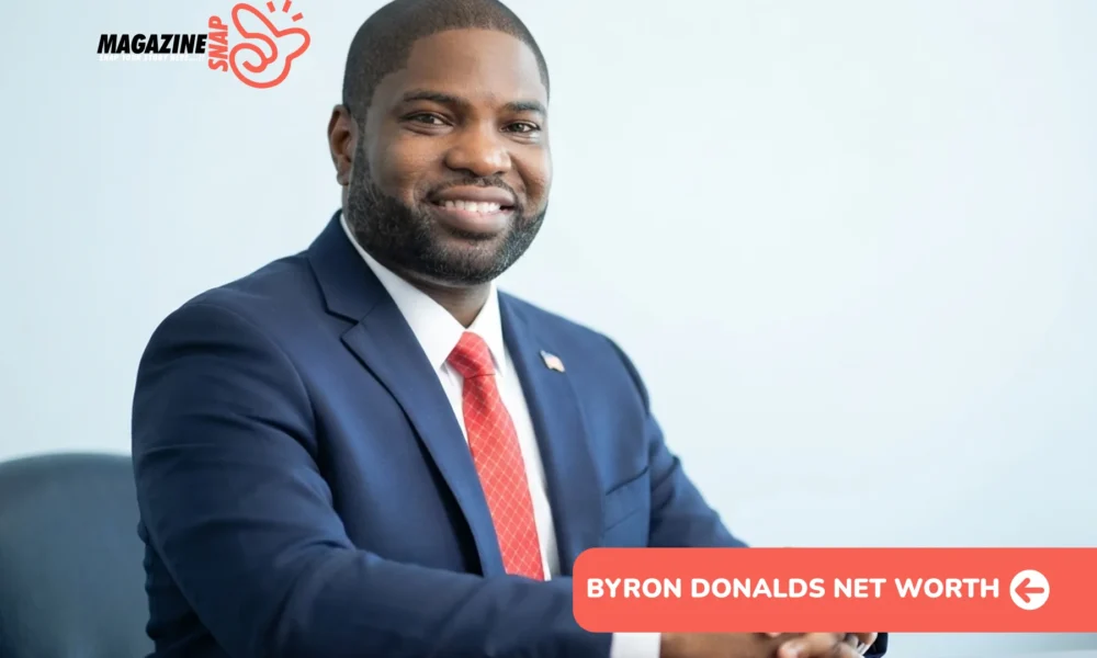 Byron Donalds smiling in navy suit and red tie, seated at wooden desk