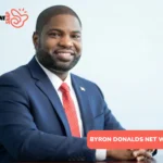 Byron Donalds smiling in navy suit and red tie, seated at wooden desk