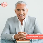 Cesar Millan Net Worth - gray-haired man in gray suit smiling at glass desk with notebook