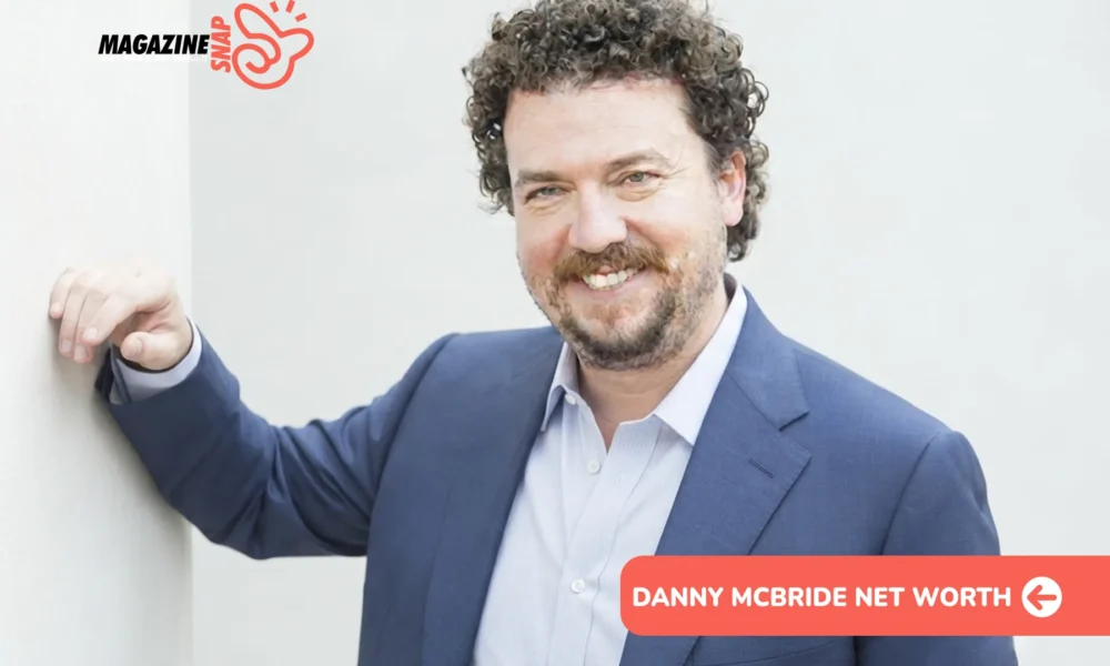 Danny McBride net worth photo: actor smiling in navy blazer, curly hair, beard