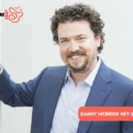 Danny McBride net worth photo: actor smiling in navy blazer, curly hair, beard