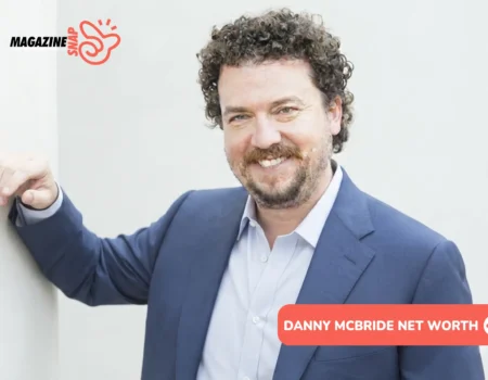 Danny McBride net worth photo: actor smiling in navy blazer, curly hair, beard