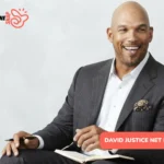 David Justice net worth portrait, smiling in gray pinstripe suit holding pen over notebook
