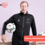 Duncan Ferguson in black Everton tracksuit holding a match ball beside a wooden table