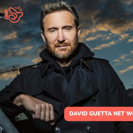 David Guetta net worth leans on a rooftop railing at dusk in a dark coat for the article.