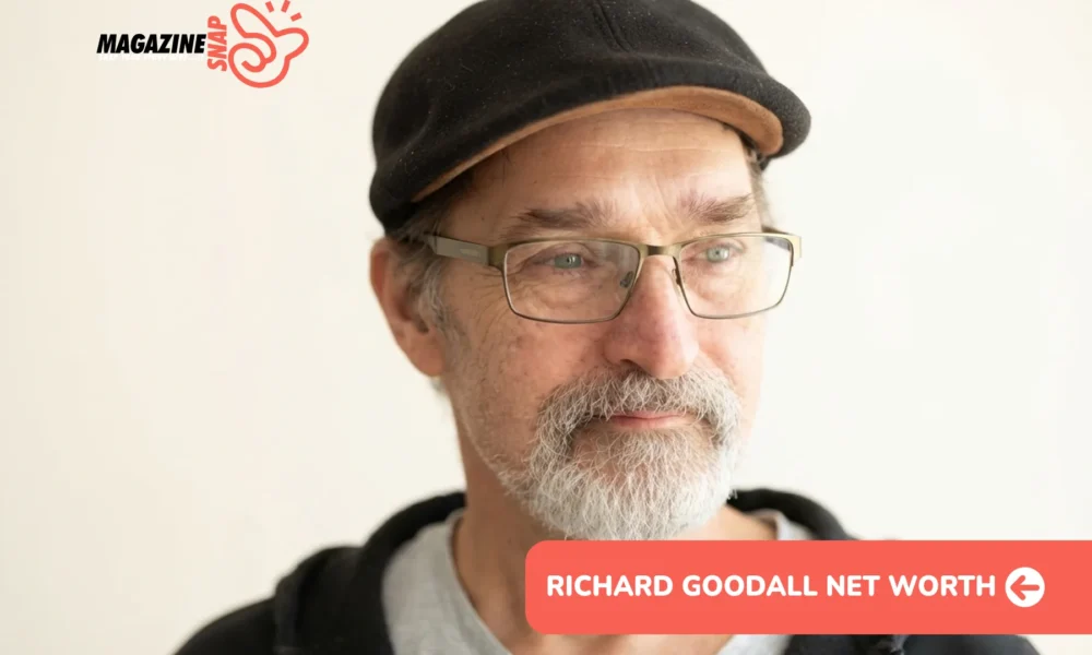 Richard Goodall net worth portrait: close-up in flat cap, glasses, and gray beard against plain background