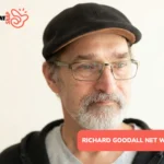 Richard Goodall net worth portrait: close-up in flat cap, glasses, and gray beard against plain background