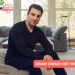 Airbnb CEO Brian Chesky sitting on a grey sofa in a bright living room, wearing a black long-sleeve shirt
