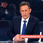 Brian Kilmeade seated at news desk in navy suit for net worth article