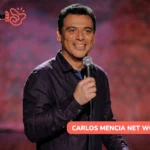 Carlos Mencia smiling on stage with a microphone in a purple shirt, Carlos Mencia net worth article photo