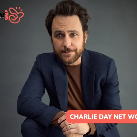 Charlie Day net worth article portrait of a bearded man in a navy blazer against a plain gray backdrop