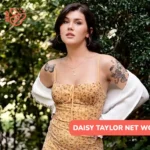 Daisy Taylor net worth article image: yellow floral dress, white cardigan, hands on hips among shrubs